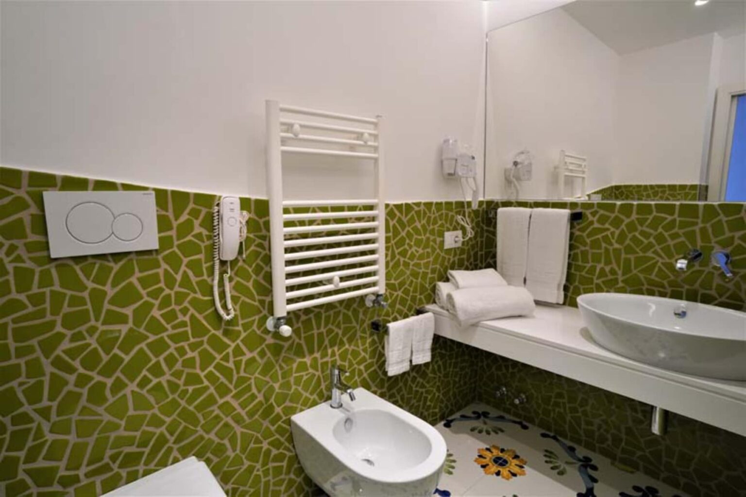 Modern bathroom design featuring green mosaic tiles, a white countertop with a round sink, a bidet, and heated towel rack. Clean and minimalist aesthetic with essential bathroom amenities. | Only on SopranoVillas