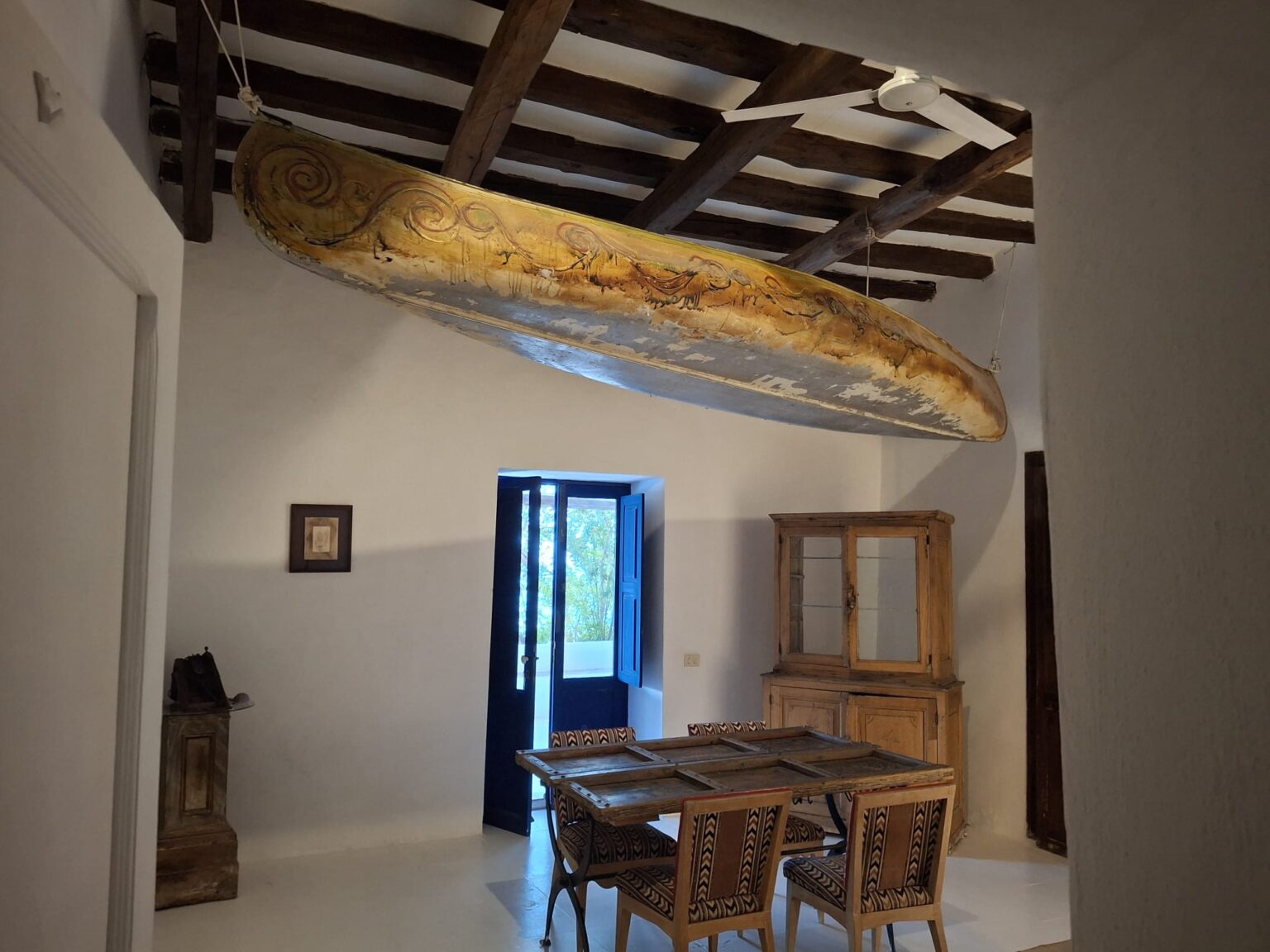 Interior view of a rustic dining area featuring a vintage boat suspended from the ceiling, wooden beams, a wooden dining table with patterned chairs, and an open doorway leading to a garden. | Only on SopranoVillas