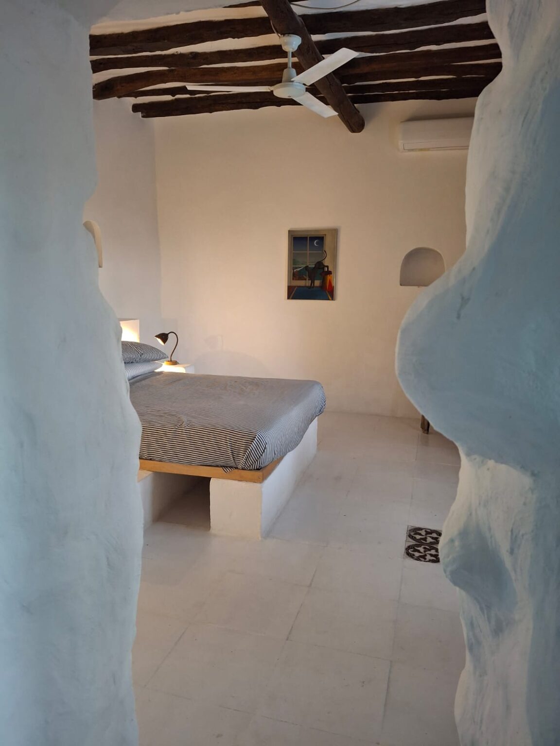 Cozy bedroom interior featuring a minimalist design with a whitewashed wall, wooden beams, and a ceiling fan. The room includes a bed with striped bedding, a bedside lamp, and an artwork on the wall, creating a serene and inviting atmosphere. Ideal for relaxation and comfort. | Only on SopranoVillas