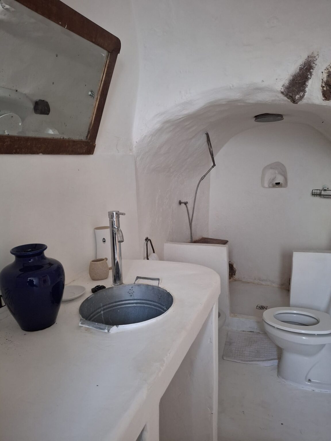 Modern bathroom interior with whitewashed walls, featuring a round sink with a metal basin, a stylish faucet, and a large mirror. The space includes a toilet and a shower area, complemented by decorative pottery on the counter. Natural light enhances the clean, minimalist design. | Only on SopranoVillas