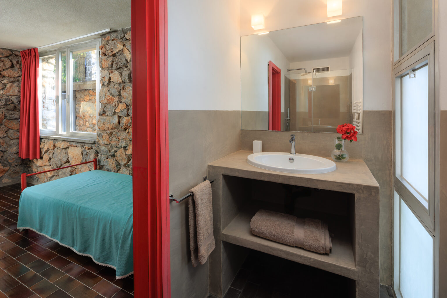 Modern bathroom with a minimalist design featuring a round sink, stone wall accents, and a cozy bedroom visible in the background with a teal bedspread and red curtains. | Only on SopranoVillas