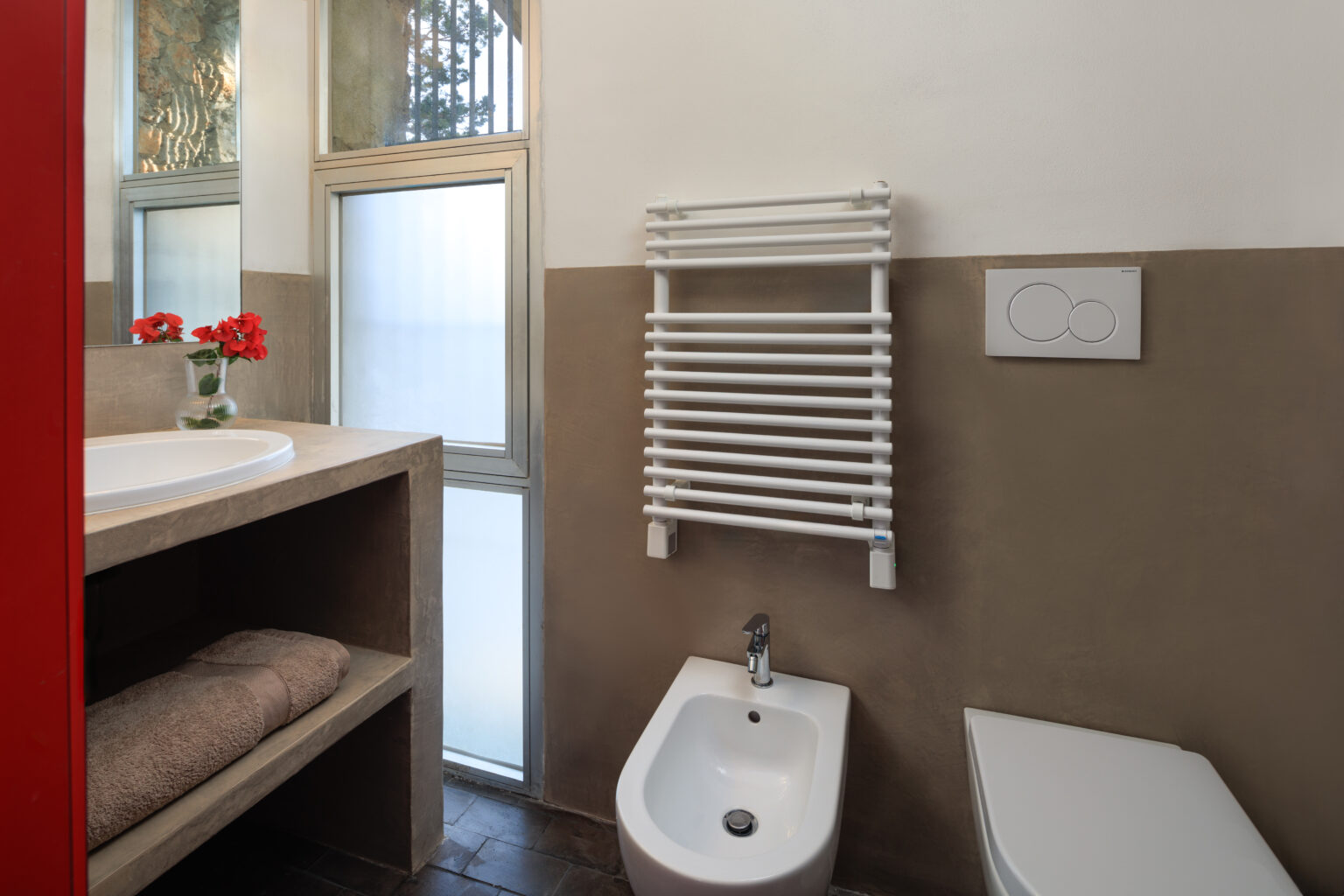 Modern bathroom interior featuring a bidet, towel rack, and a minimalist design with neutral color palette. Natural light enters through a window, complemented by decorative flowers on the countertop. | Only on SopranoVillas