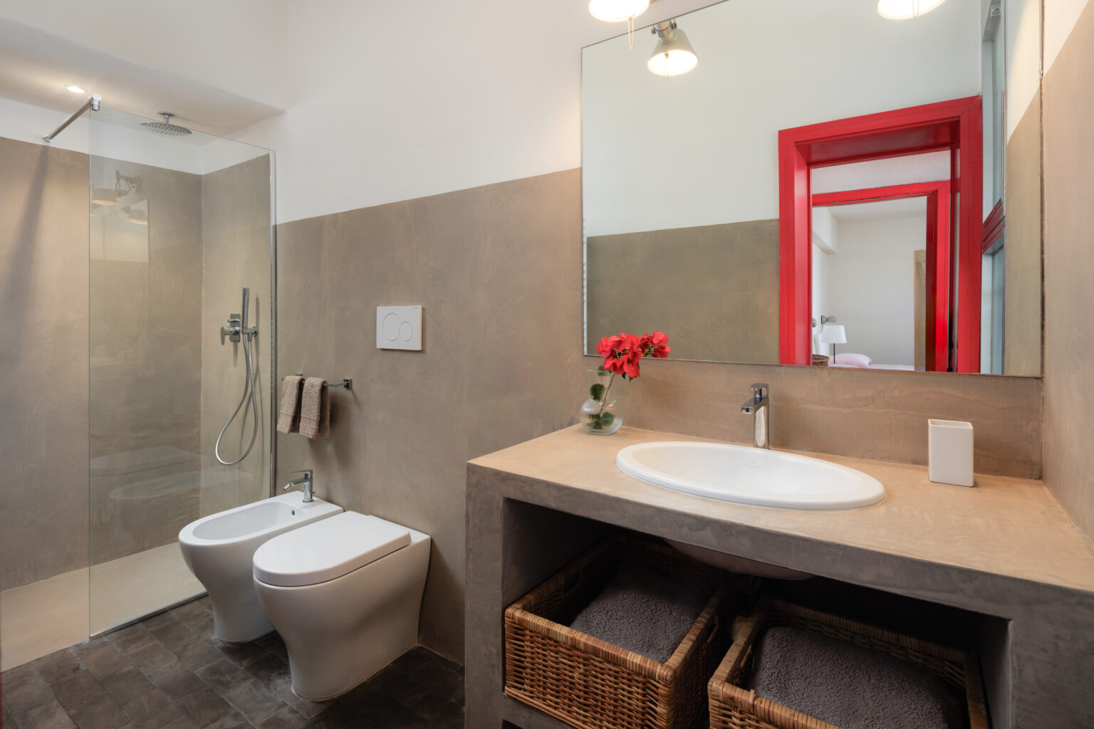 Modern bathroom featuring a glass shower, bidet, and stylish sink with a minimalist design. The decor includes a vibrant red mirror frame and a vase with flowers, complemented by woven baskets for storage. | Only on SopranoVillas