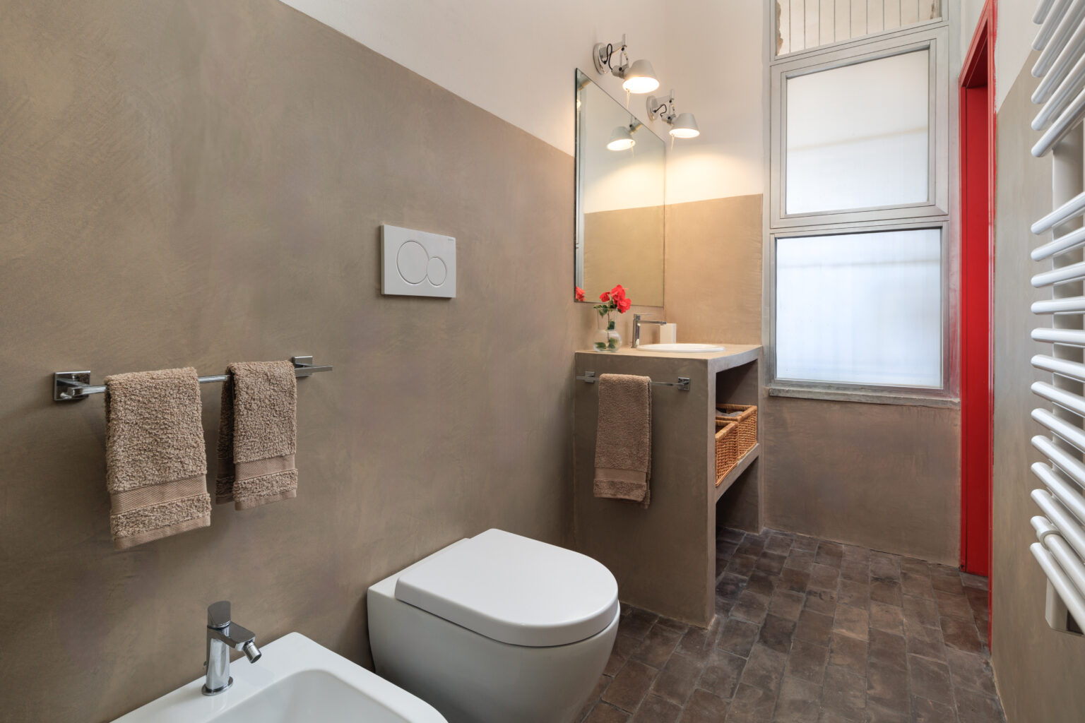 Modern bathroom interior featuring a sleek toilet, a stylish sink with storage, and decorative towels. The walls have a warm, textured finish, complemented by natural light from a window. | Only on SopranoVillas