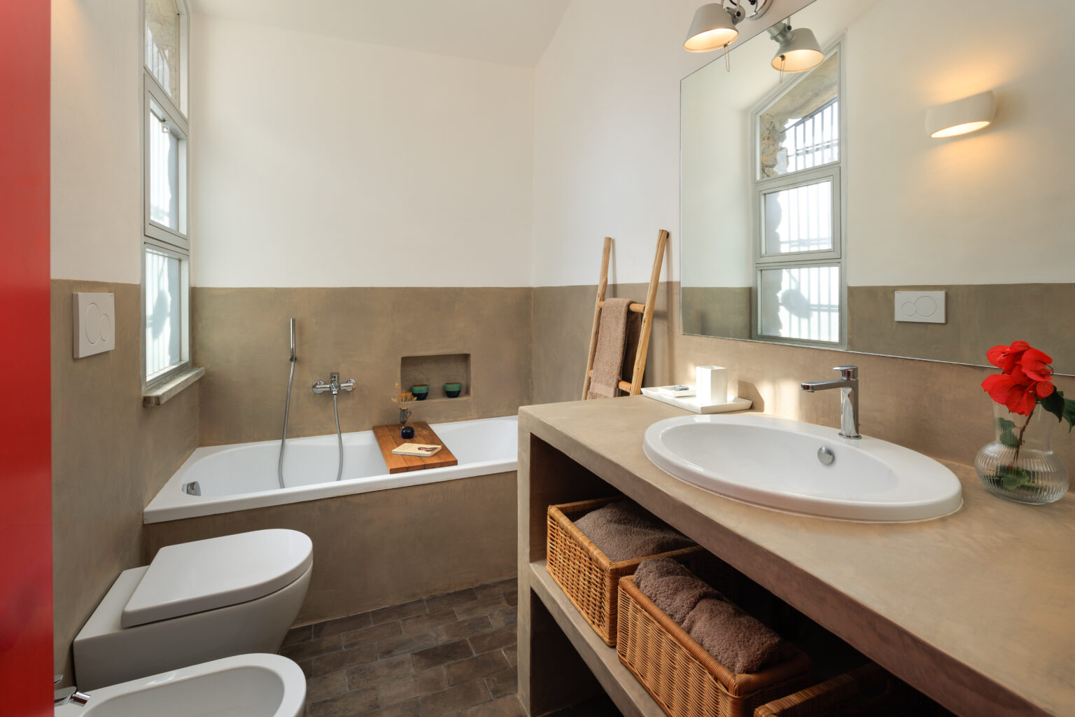 Modern bathroom interior featuring a freestanding bathtub, minimalist design, and natural light. The space includes a stylish vanity with a round sink, wooden accents, and decorative elements like flowers. The walls have a neutral color palette, complemented by stone flooring and a wooden ladder for towel storage, creating a serene atmosphere. | Only on SopranoVillas