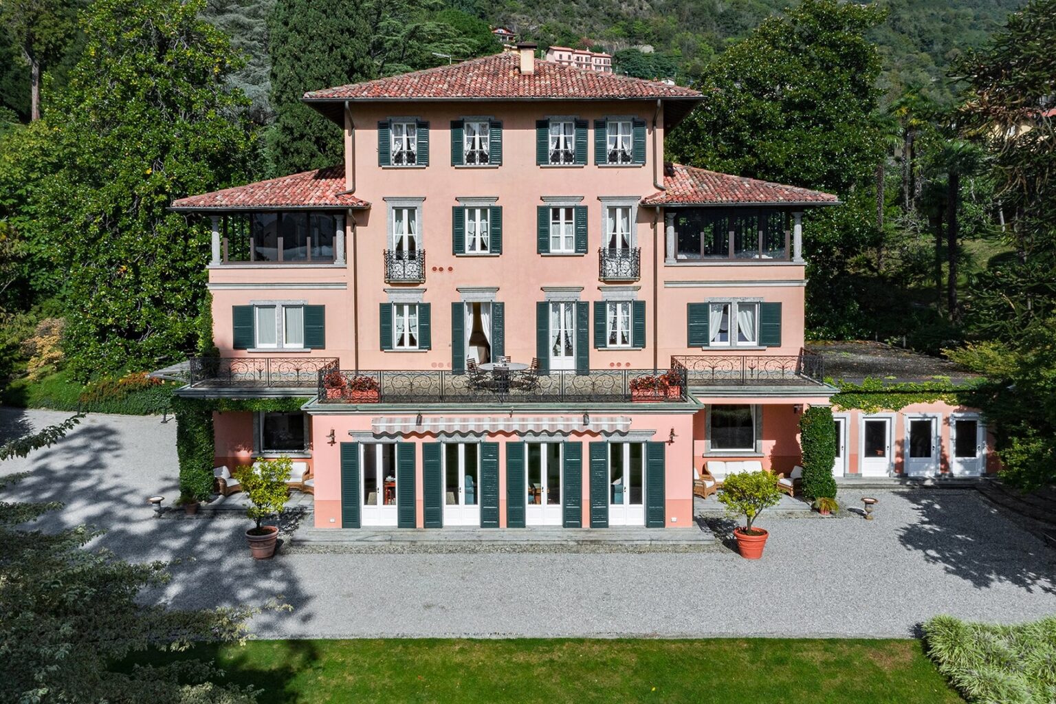 Aerial view of a charming pink villa with a red-tiled roof, featuring multiple balconies, green shutters, and a spacious gravel driveway surrounded by lush greenery. The villa showcases elegant architecture and well-maintained gardens, ideal for luxury living or vacation rentals. | Only on SopranoVillas