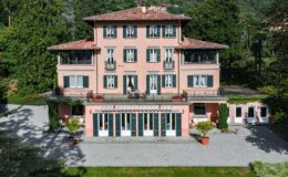 Aerial view of a charming pink villa with a red-tiled roof, featuring multiple balconies, green shutters, and a spacious gravel driveway surrounded by lush greenery. The villa showcases elegant architecture and well-maintained gardens, ideal for luxury living or vacation rentals.