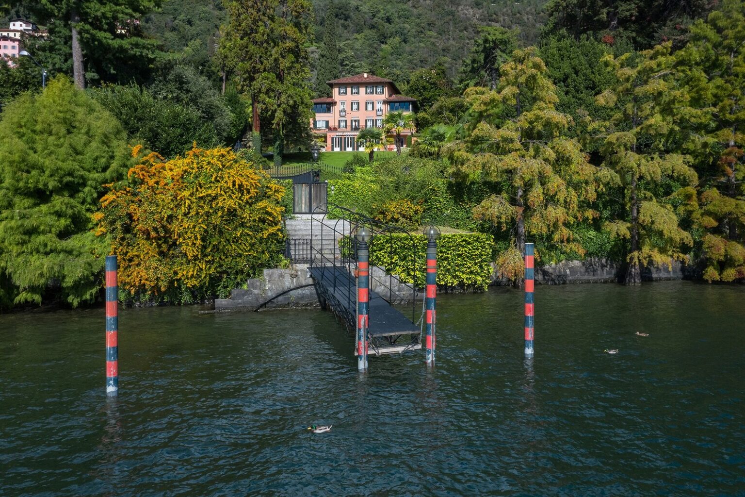Scenic view of a lakeside property with a charming house surrounded by lush greenery and colorful trees, featuring a wooden dock extending into the water. | Only on SopranoVillas