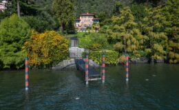 Scenic view of a lakeside property with a charming house surrounded by lush greenery and colorful trees, featuring a wooden dock extending into the water.
