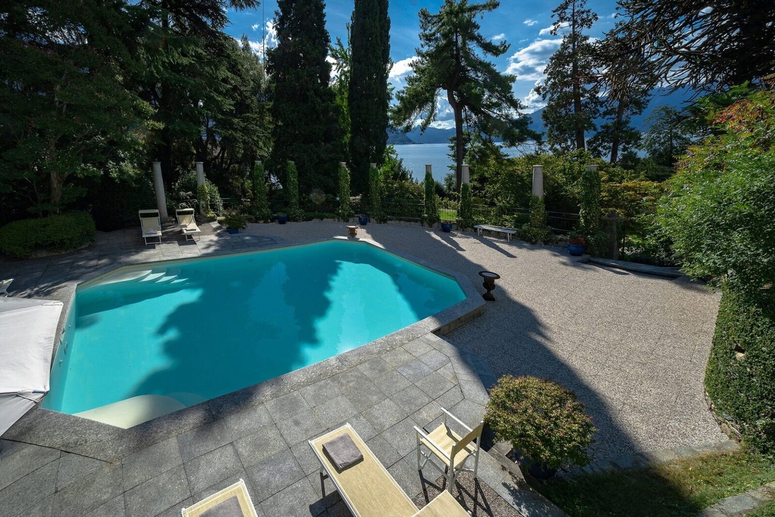 Scenic view of a luxurious outdoor swimming pool surrounded by lush greenery and tall trees, with a tranquil lake in the background under a clear blue sky. | Only on SopranoVillas