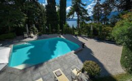 Scenic view of a luxurious outdoor swimming pool surrounded by lush greenery and tall trees, with a tranquil lake in the background under a clear blue sky.