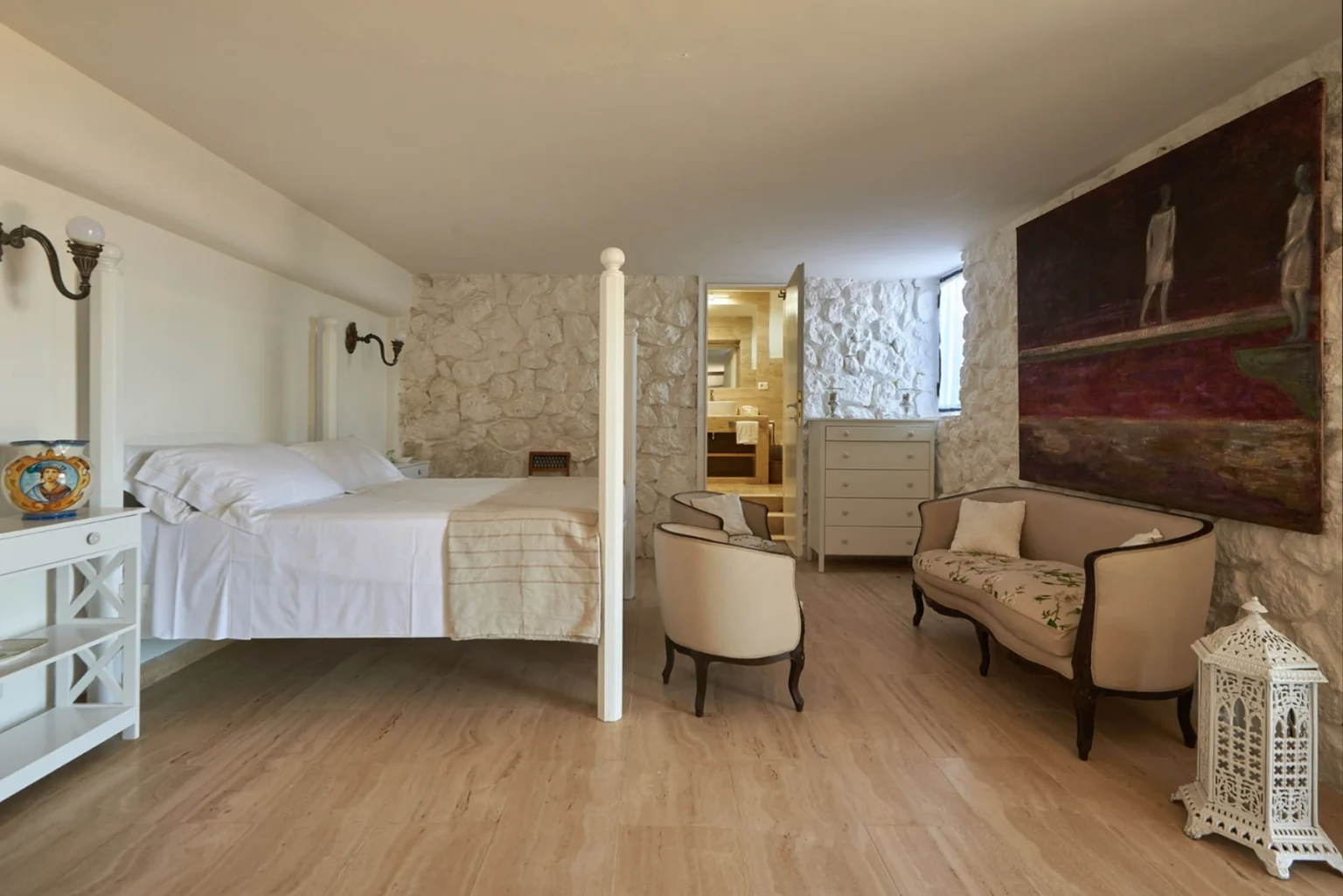 Cozy bedroom featuring a four-poster bed with white linens, stone accent walls, and elegant furnishings, including a vintage-style armchair and a decorative lantern. The room also includes a dresser and leads to a modern bathroom, creating a blend of rustic charm and contemporary design. | Only on SopranoVillas