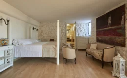 Cozy bedroom featuring a four-poster bed with white linens, stone accent walls, and elegant furnishings, including a vintage-style armchair and a decorative lantern. The room also includes a dresser and leads to a modern bathroom, creating a blend of rustic charm and contemporary design.