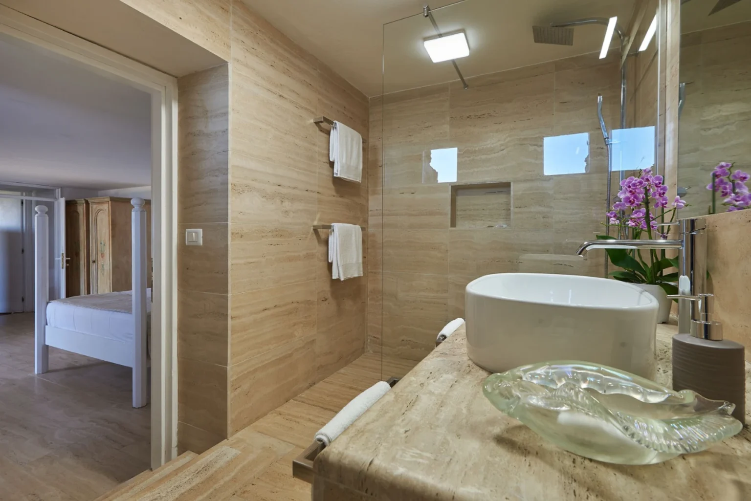 Modern bathroom design featuring natural stone walls, a glass shower enclosure, and a stylish countertop with a white sink. The decor includes fresh orchids and neatly arranged towels, creating a serene and elegant atmosphere. | Only on SopranoVillas