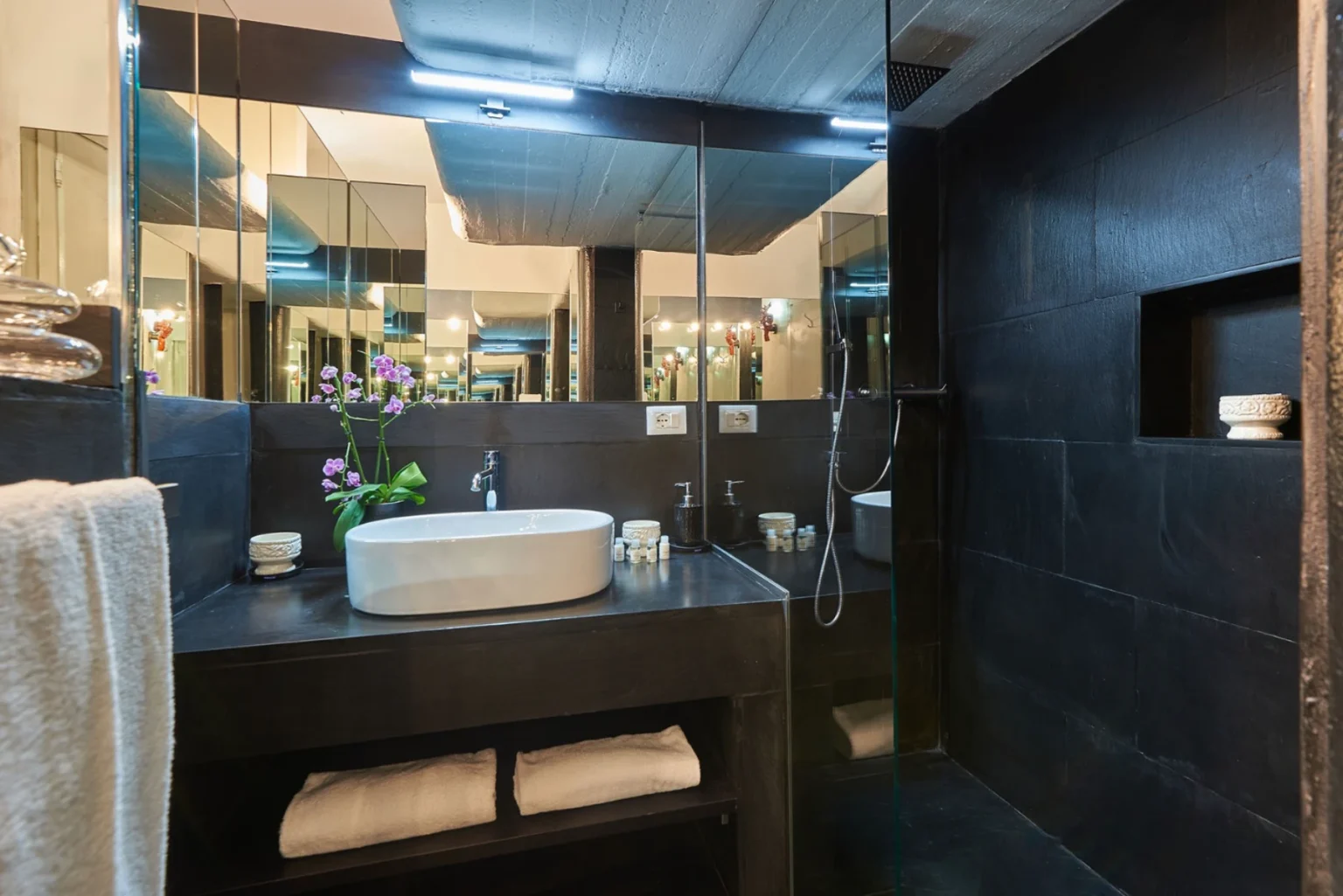 Modern bathroom interior featuring a sleek black stone design, a white oval sink, decorative orchid plant, and neatly arranged towels, with ample mirrors reflecting light for a spacious feel. Ideal for luxury home decor inspiration. | Only on SopranoVillas