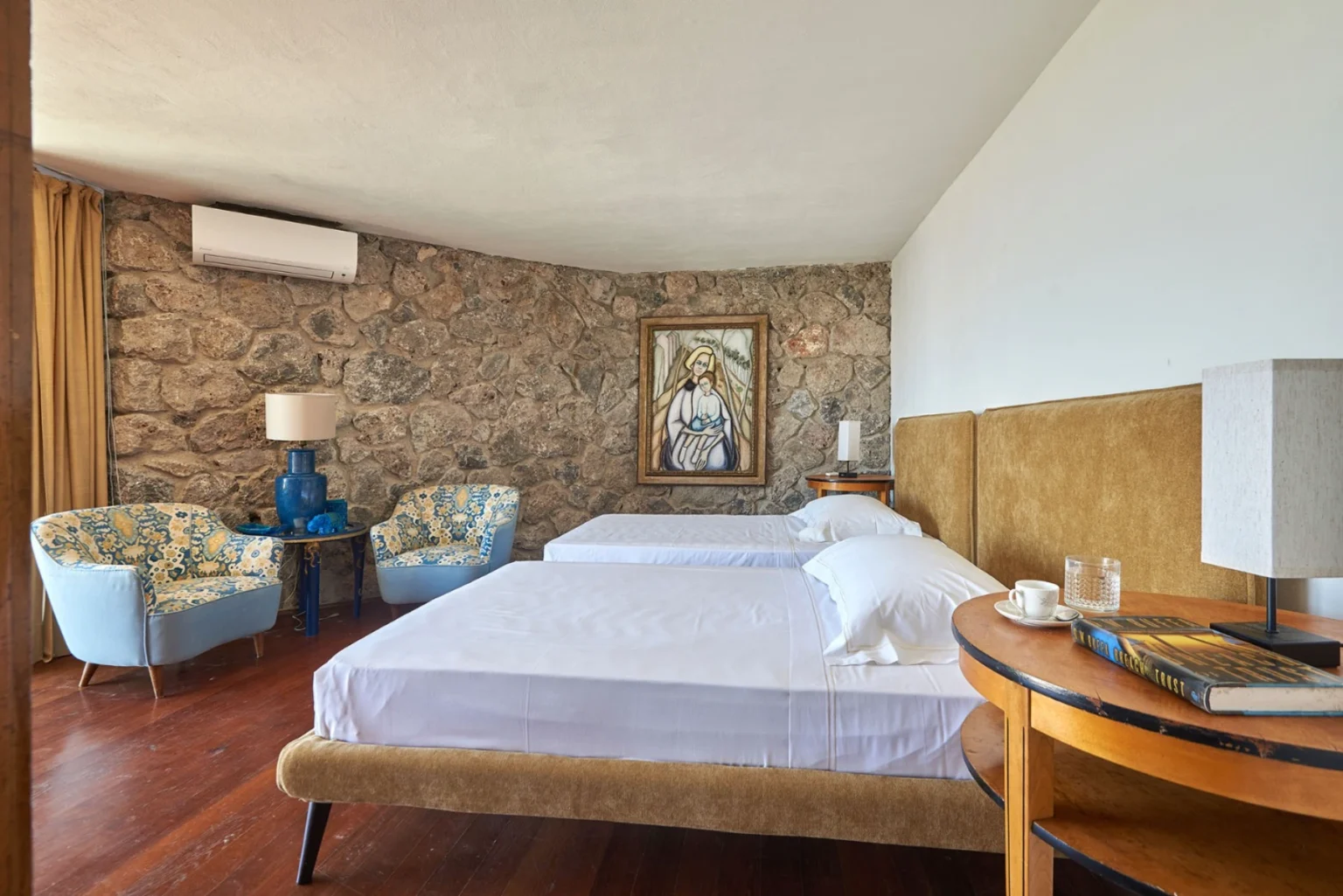 Cozy hotel room featuring two twin beds, a stone wall, and stylish blue-and-yellow armchairs. Decor includes a religious painting, bedside tables with lamps, and a coffee cup on a wooden table, creating a warm and inviting atmosphere. | Only on SopranoVillas