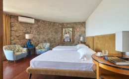 Cozy hotel room featuring two twin beds, a stone wall, and stylish blue-and-yellow armchairs. Decor includes a religious painting, bedside tables with lamps, and a coffee cup on a wooden table, creating a warm and inviting atmosphere.