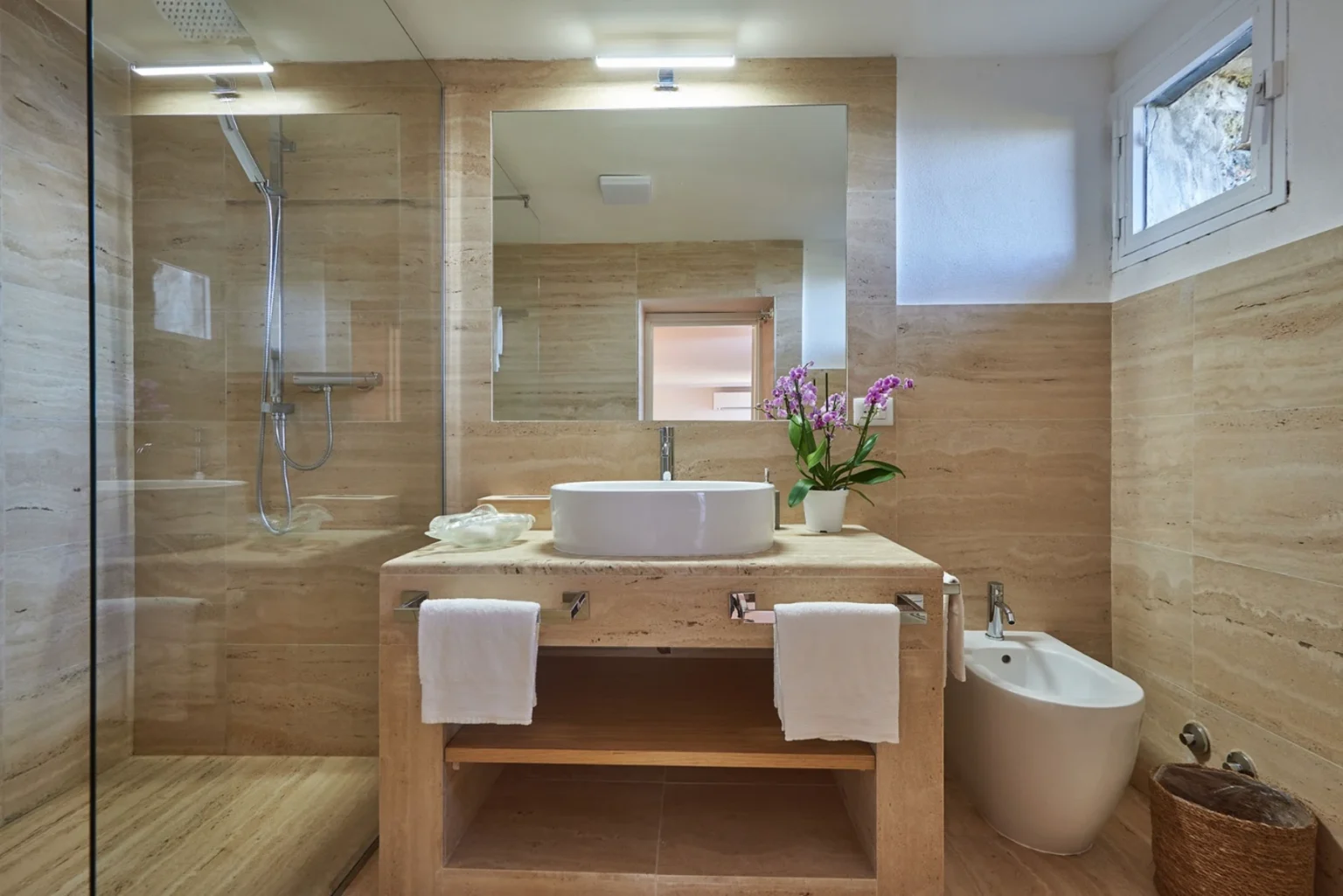 Modern bathroom featuring natural stone walls, a sleek glass shower, a stylish vessel sink on a wooden vanity, fresh orchids as decor, and elegant white towels. | Only on SopranoVillas