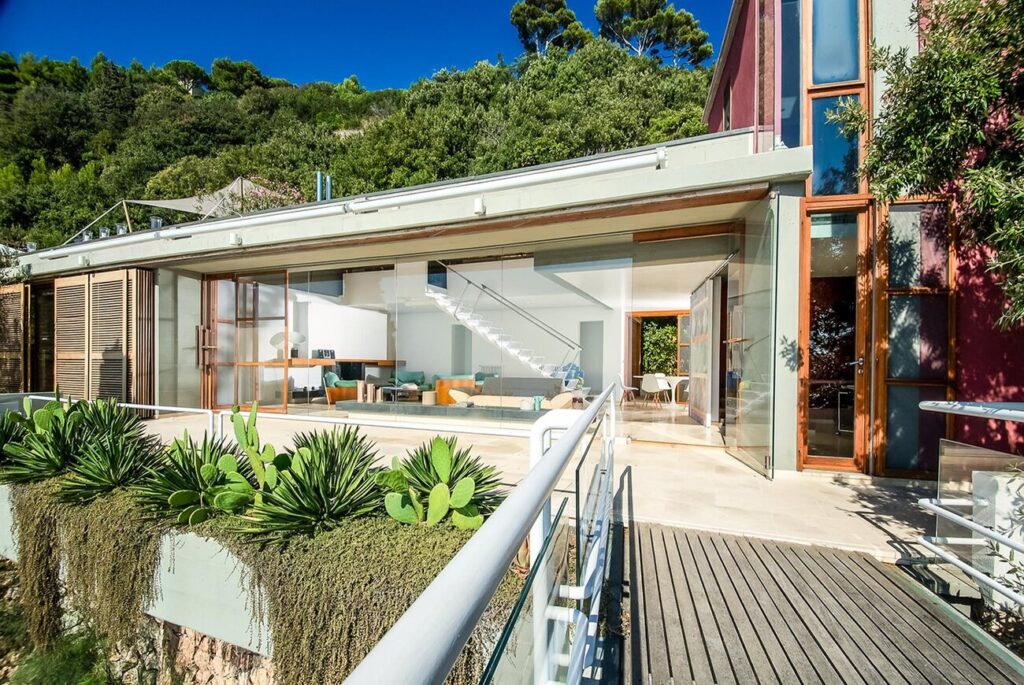 Modern eco-friendly house with large glass windows and wooden accents, featuring a landscaped deck and lush greenery in the background. Ideal for showcasing sustainable architecture and outdoor living spaces. | Only on SopranoVillas