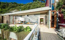 Modern eco-friendly house with large glass windows and wooden accents, featuring a landscaped deck and lush greenery in the background. Ideal for showcasing sustainable architecture and outdoor living spaces.