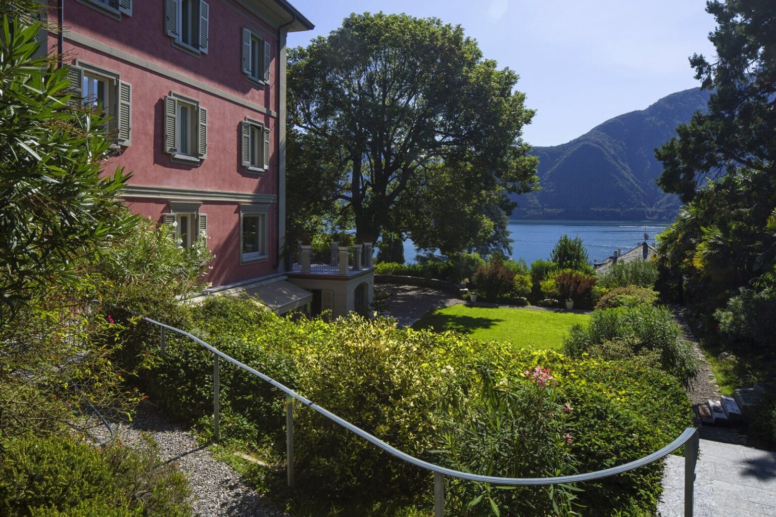 Charming pink villa surrounded by lush greenery and a serene lake view, featuring a well-maintained garden and a pathway leading to the water, set against a backdrop of mountains under a clear blue sky. | Only on SopranoVillas