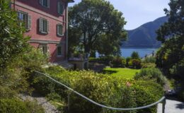 Charming pink villa surrounded by lush greenery and a serene lake view, featuring a well-maintained garden and a pathway leading to the water, set against a backdrop of mountains under a clear blue sky.