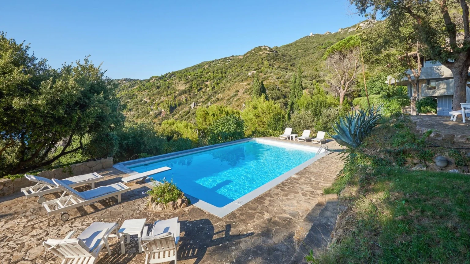 Scenic outdoor swimming pool surrounded by lush greenery and mountains, featuring white lounge chairs and a stone patio, ideal for relaxation and leisure. | Only on SopranoVillas