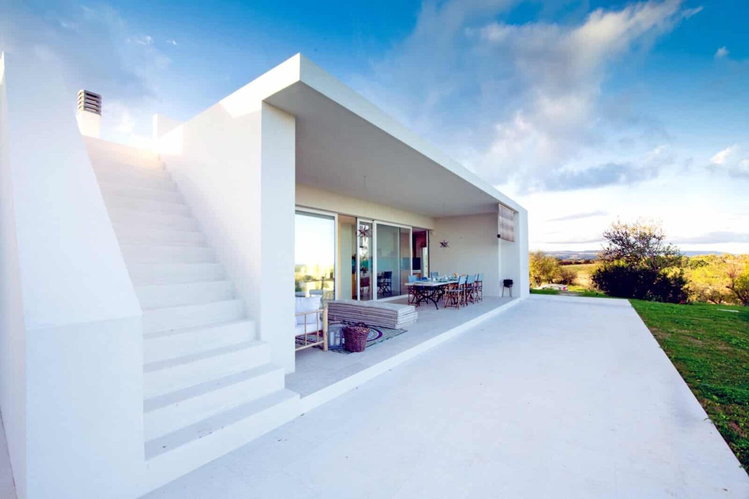 Modern white house with large glass windows and a minimalist design, featuring a staircase and a spacious outdoor area, set against a scenic landscape. | Only on SopranoVillas