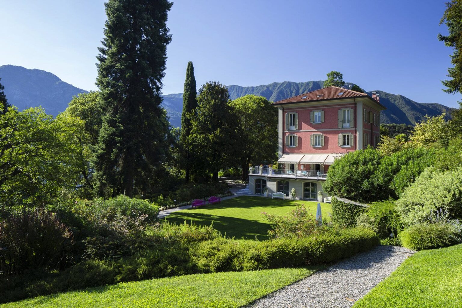 Historic villa surrounded by lush gardens and tall trees, with mountains in the background under a clear blue sky. | Only on SopranoVillas