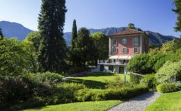 Historic villa surrounded by lush gardens and tall trees, with mountains in the background under a clear blue sky.