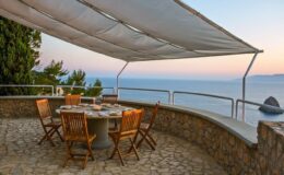 Elegant outdoor dining area with wooden chairs and a round table, set against a stunning ocean view at sunset. The area is shaded by a canopy, providing a relaxing atmosphere for meals and gatherings.