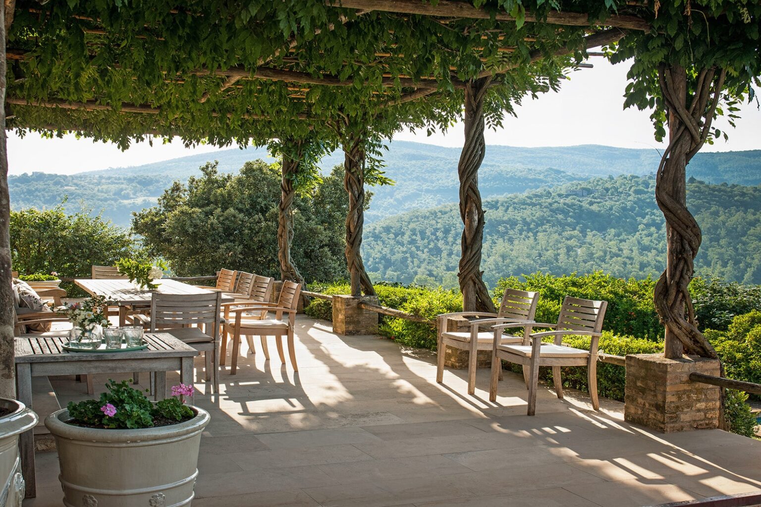 Scenic outdoor patio with wooden furniture, surrounded by lush greenery and mountains in the background, perfect for relaxation and enjoying nature. | Only on SopranoVillas