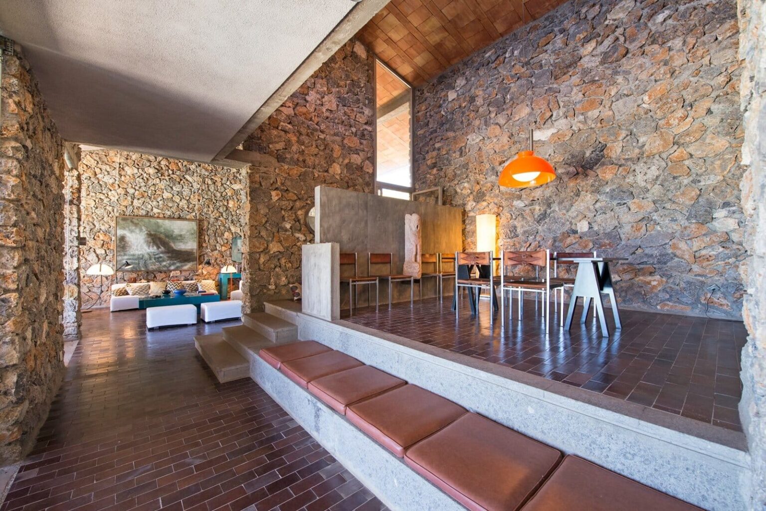 Modern living room featuring stone walls, wooden flooring, and minimalist furniture. The space includes a raised seating area with brown cushions, a dining table, and an orange pendant light, creating a warm and inviting atmosphere. | Only on SopranoVillas