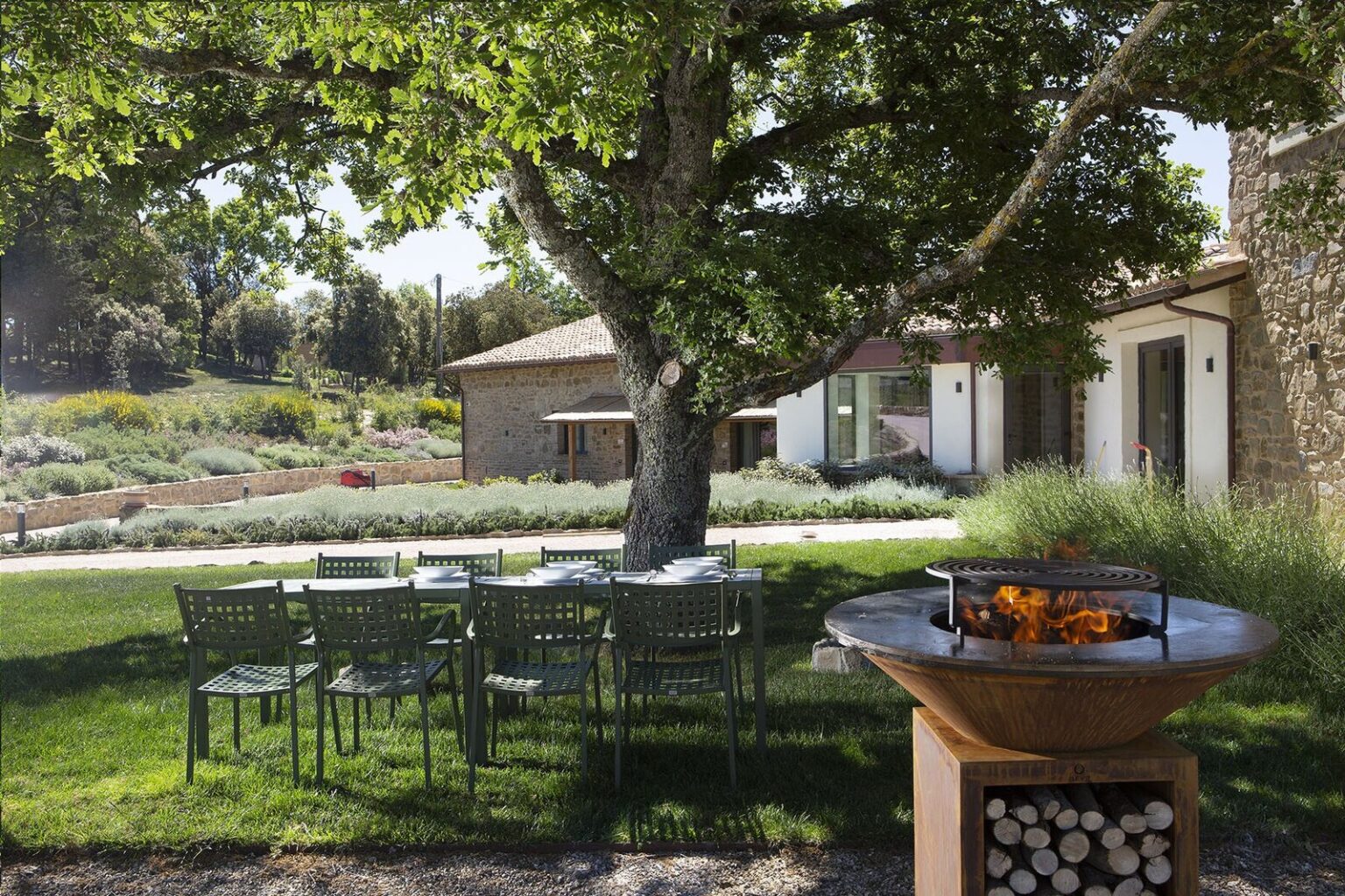 Outdoor dining area with green chairs and a fire pit under a large tree, featuring a scenic background of a landscaped garden and modern architecture. | Only on SopranoVillas