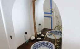 Elegant bathroom setting featuring a wooden mirror frame, ornate sink with blue and white ceramic design, and minimalistic decor. The wall is painted white with a blue accent door, creating a serene and stylish atmosphere.