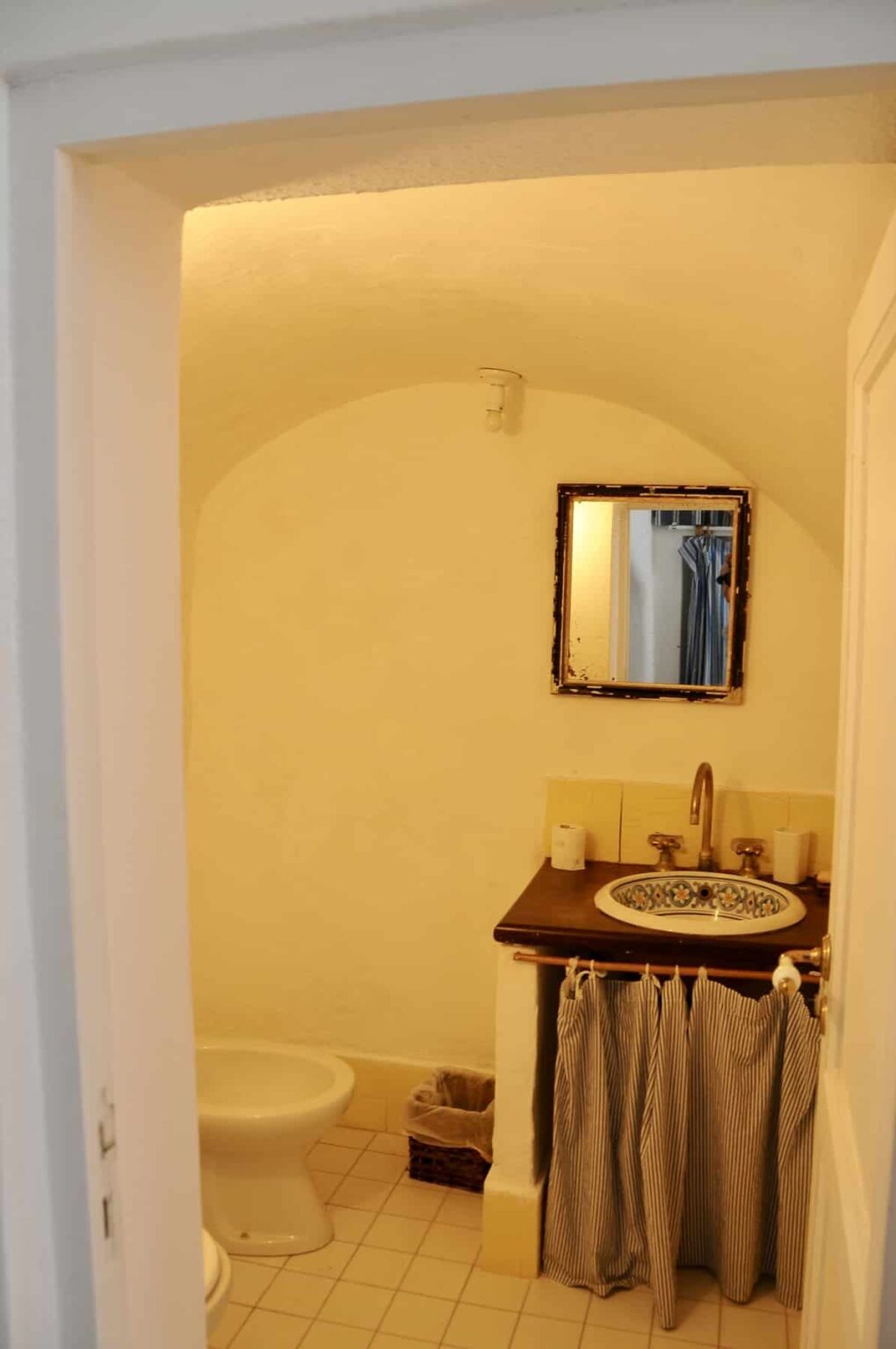 Small bathroom with a yellow stucco wall, featuring a vintage mirror above a sink with decorative tiles, and a wooden countertop. | Only on SopranoVillas