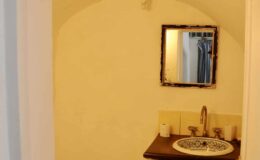 Small bathroom with a yellow stucco wall, featuring a vintage mirror above a sink with decorative tiles, and a wooden countertop.