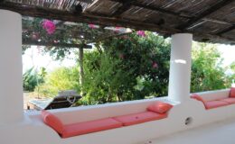 Cozy outdoor seating area with orange cushions under a thatched roof, surrounded by lush greenery and blooming flowers, ideal for relaxation and enjoying nature.