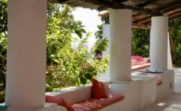 Cozy outdoor seating area with white columns, surrounded by lush greenery and vibrant flowers, offering a tranquil atmosphere for relaxation.