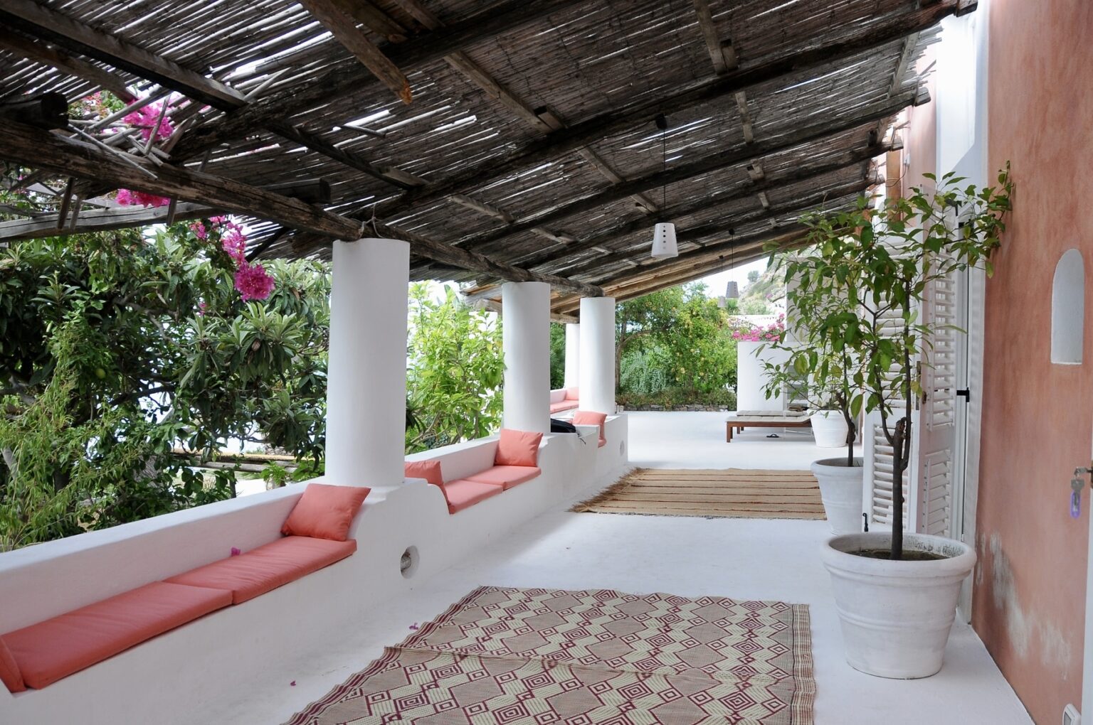 Cozy outdoor terrace with pink cushioned seating, surrounded by greenery and a decorative rug, featuring a wooden overhead canopy for shade. | Only on SopranoVillas