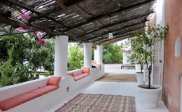 Cozy outdoor terrace with pink cushioned seating, surrounded by greenery and a decorative rug, featuring a wooden overhead canopy for shade.