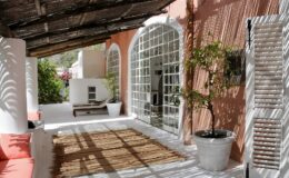 Bright and airy outdoor patio with terracotta walls, white columns, and a thatched roof, featuring a woven rug, potted plants, and a comfortable seating area, ideal for relaxation and enjoying nature.