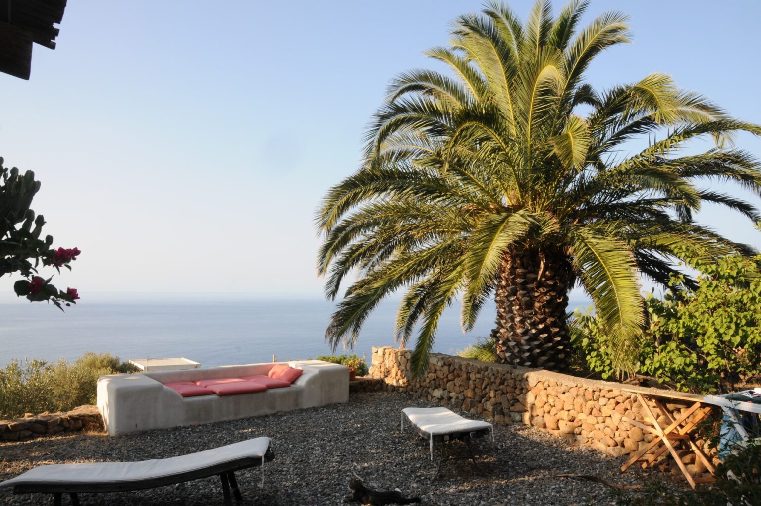 Scenic outdoor space featuring a large palm tree, comfortable seating area with pink cushions, and a view of the ocean. The setting includes gravel ground and stone walls, creating a serene atmosphere ideal for relaxation. | Only on SopranoVillas