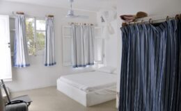 Bright and airy bedroom featuring a white bed, light-colored walls, and blue-striped curtains. The room includes a ceiling fan and a simple seating area, creating a minimalist and inviting atmosphere. Ideal for relaxation and comfort.