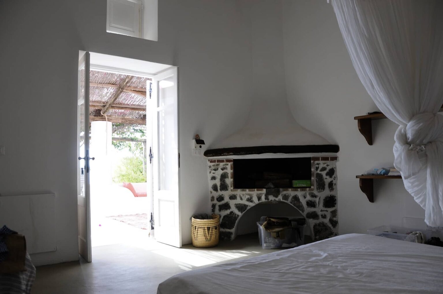Cozy bedroom interior featuring a stone fireplace, white walls, and an open doorway leading to an outdoor patio with natural light streaming in. A woven basket and shelves are visible, enhancing the inviting atmosphere. | Only on SopranoVillas