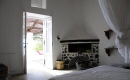 Cozy bedroom interior featuring a stone fireplace, white walls, and an open doorway leading to an outdoor patio with natural light streaming in. A woven basket and shelves are visible, enhancing the inviting atmosphere.