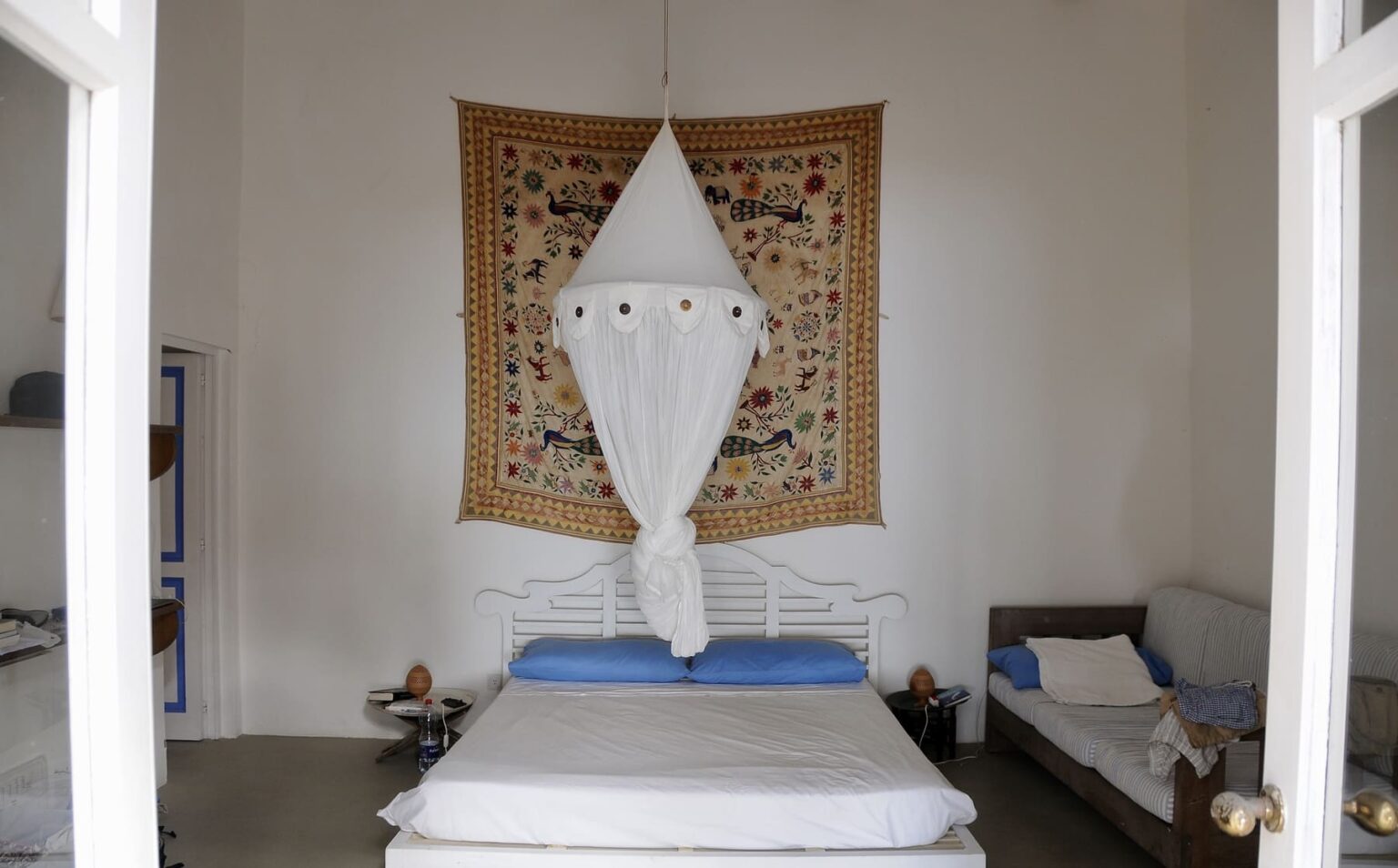Cozy bedroom featuring a white bed with blue pillows, adorned with a decorative canopy, and a colorful wall tapestry. The room has a minimalist design with light walls and a wooden sofa, creating a serene atmosphere. | Only on SopranoVillas