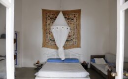 Cozy bedroom featuring a white bed with blue pillows, adorned with a decorative canopy, and a colorful wall tapestry. The room has a minimalist design with light walls and a wooden sofa, creating a serene atmosphere.