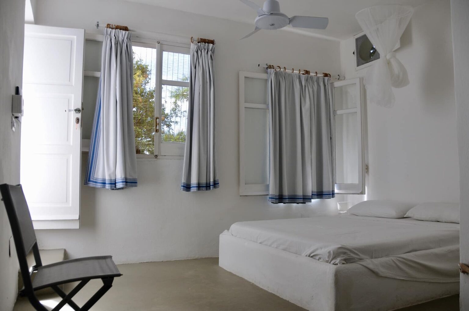 Bright and airy bedroom featuring a white bed, light-colored walls, and large windows with blue and white curtains, providing a serene and inviting atmosphere. | Only on SopranoVillas
