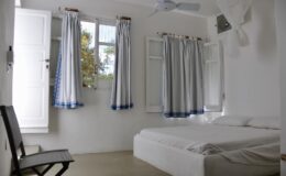 Bright and airy bedroom featuring a white bed, light-colored walls, and large windows with blue and white curtains, providing a serene and inviting atmosphere.